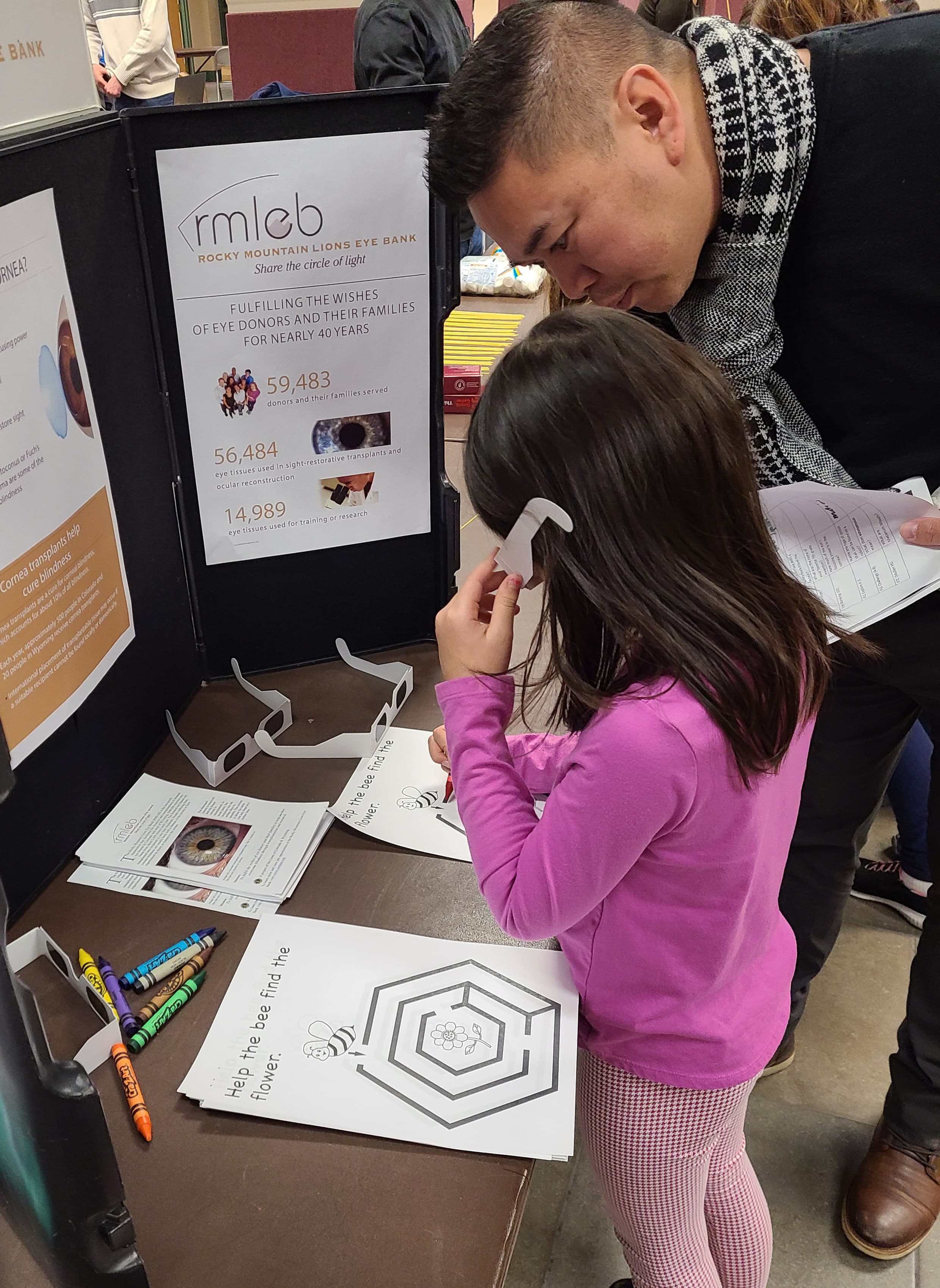 A father and daughter at an RMLEB education table, the girl trying on blindness-simulation glasses.